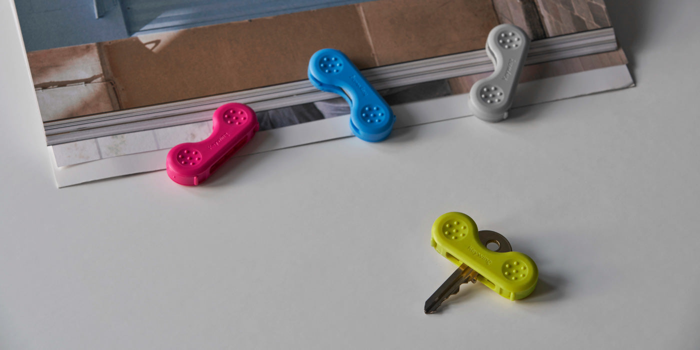 Four keywing key turner aids on a table in pink, blue, grey, yellow. Clipped onto a key to give extra leverage for weak grip.