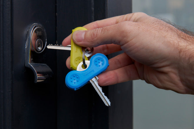 A green and blue keywing key turner aid on a keyring held by a hand that is unlocking a dark blue door with silver lock.